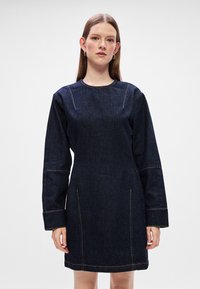 Σκούρο μπλε τζιν φόρεμα με μακριά μανίκια, στρογγυλή λαιμόκοψη και εμφανείς ραφές. Διαθέτει δύο πλαϊνές τσέπες μπροστά.