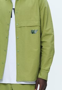 Light green button-up jacket with a large chest pocket and printed logo. Smooth texture and snap buttons dot the front, featuring a relaxed fit.