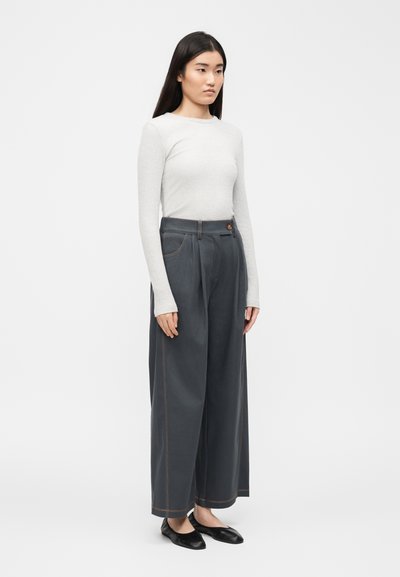 Νεαρή γυναίκα που φορά γκρι φαρδιά παντελόνια, άσπρη μακρυμάνικη ριμπ μπλούζα και μαύρα επίπεδα παπούτσια, στέκεται μπροστά σε απλό λευκό φόντο.