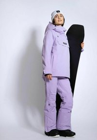 Lavendel snowboardjakke og -bukser har vandtæt materiale, en forlomma og en afslappet pasform. En sort snowboard holdes ind mod kroppen.