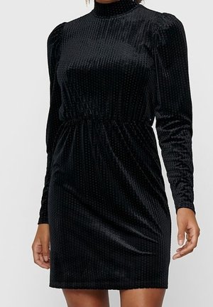 Czarna aksamitna sukienka z długimi bufiastymi rękawami, wysokim kołnierzem i marszczoną talią, noszona przez stojącą osobę.