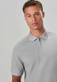 Graues kurzärmeliges Polohemd mit Reißverschluss am Kragen und strukturiertem Stoff. Das Design ist leger mit lockerem Sitz und dezenten Details.