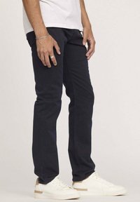 Pantalons en coton bleu marine avec une coupe droite, couture de poche discrète, associés à des baskets blanches. Le tissu présente une texture lisse et une durabilité.
