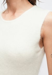 Närbild av en person som bär en ärmlös, fluffig vit topp, där halsen, axeln och en del av en tatuering på överarmen syns.