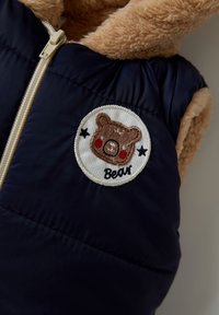 Marineblaues Puffer-Weste mit kragen aus beigefarbenem Kunstfell, ausgestattet mit einem runden gestickten Patch eines Bären mit roten Wangen und dem Wort "Bär".