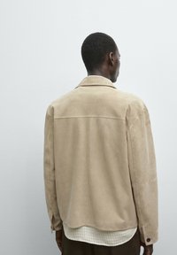 Beige Wildlederjacke mit einem entspannten Schnitt, weitem Kragen und Rückenpasse. Darunter eine karierte Bluse mit abgerundetem Saum und dunklen Hosen.