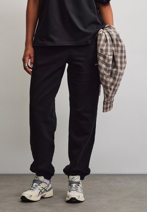 Persona con pantaloni neri e scarpe da ginnastica che tiene in mano una camicia beige e nera a quadri piegata su uno sfondo grigio uniforme.