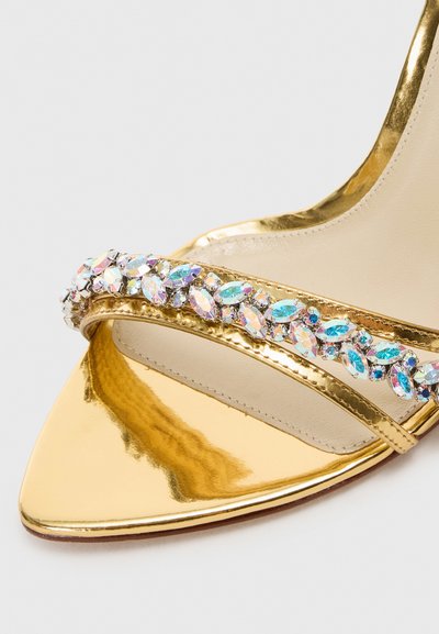 Sandalo con tacco alto dorato, caratterizzato da punta affusolata, cinturino metallico e decorato con pietre preziose sfaccettate e colorate lungo il cinturino anteriore.