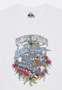 Witte t-shirt met een kleurrafisch van een skelet dat surft op een golf, omringd door tropische planten en een geruit surfboard.