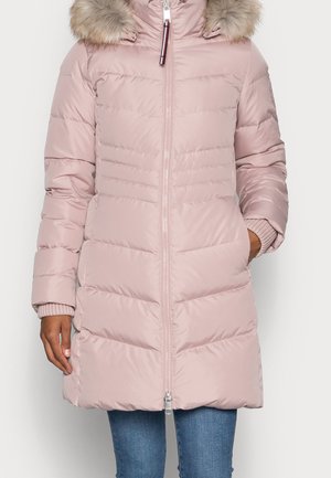 Manteau d’hiver matelassé rose clair avec une fermeture éclair à l’avant, capuche bordée de fourrure, mains dans les poches, porté avec un jean bleu.