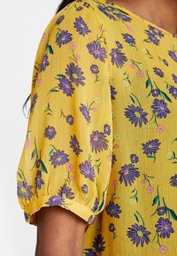 Blouse jaune à manches courtes bouffantes, arborant un motif floral de marguerites violettes et de fleurs roses sur un tissu texturé.