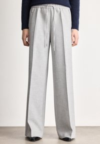 Persona valkā gaiši pelēkas, platas bikses ar auklu viduklī, kombinētas ar melniem apaviem un tumši zilu džemperi.