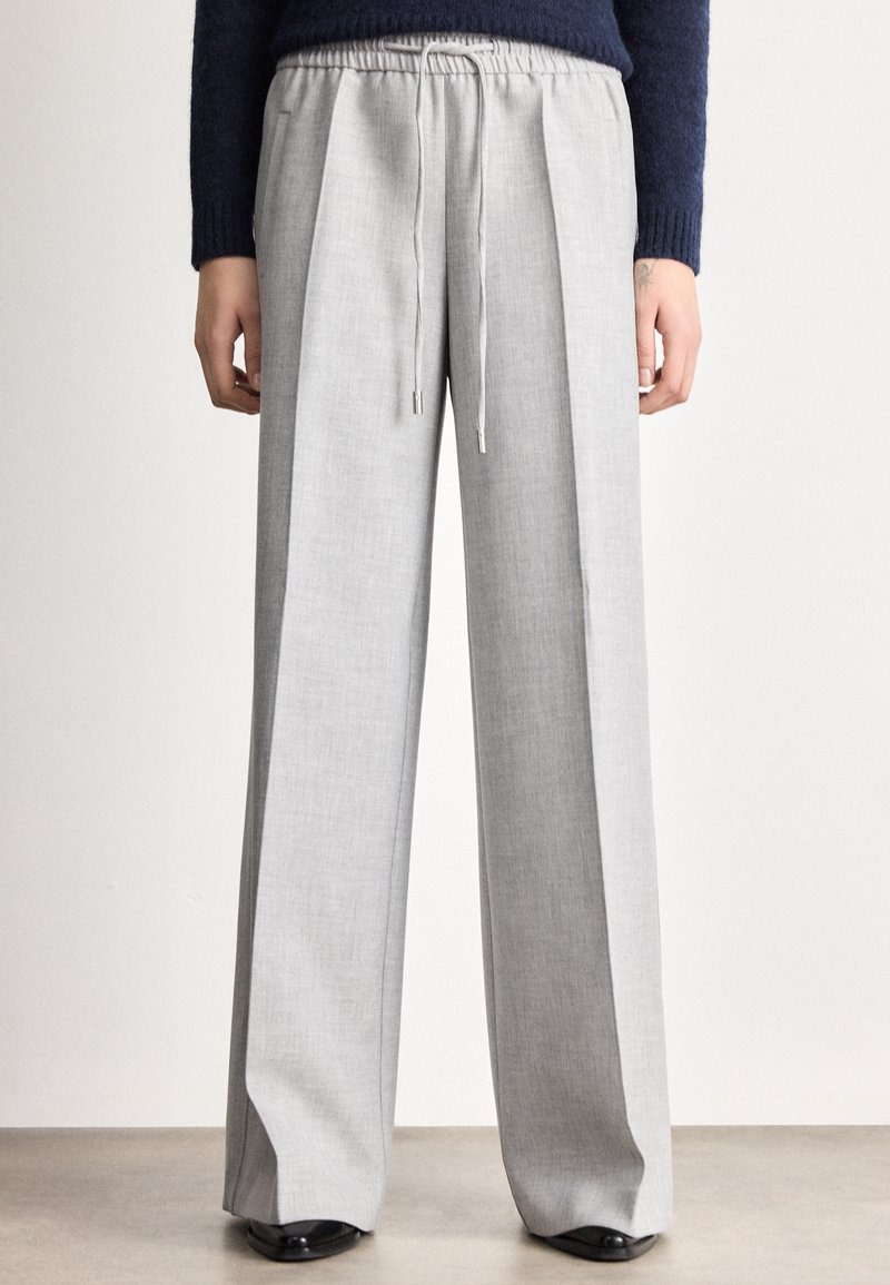 Persona valkā gaiši pelēkas, platas bikses ar auklu viduklī, kombinētas ar melniem apaviem un tumši zilu džemperi.