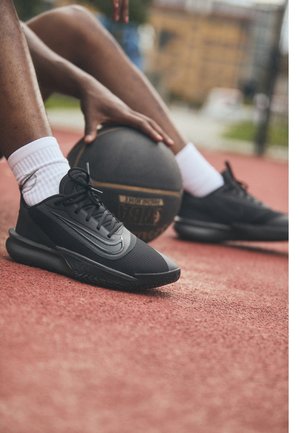 Persona con zapatillas negras y calcetines blancos sosteniendo un balón de baloncesto mientras está sentada en una cancha exterior roja.