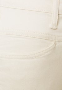 Gros plan sur un tissu en denim blanc cassé montrant une poche arrière avec des coutures détaillées et une passoire de ceinture.