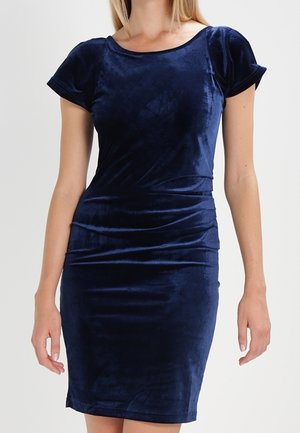 Femme portant une robe en velours bleu marine ajustée à manches courtes, avec un col rond et un détail froncé à la taille.