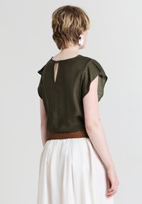 Top corto maniche corte verde oliva con dettaglio a chiave sul retro, abbinato a una cintura intrecciata marrone. Il tessuto ha una finitura morbida e testurizzata.