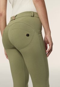 Vihreät stretch-housut, joissa on sileä pinta, kaarevat takataskut ja pieni kultainen logokoriste vasemmassa taskussa.