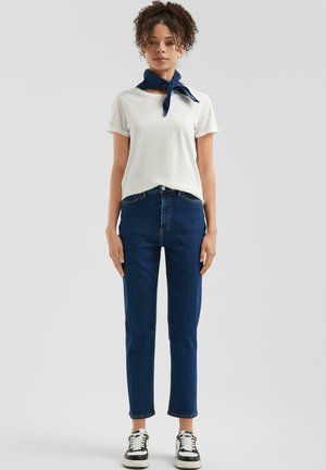 Weißes Baumwoll-T-Shirt mit hochgekrempelten Ärmeln, kombiniert mit hochtaillierten blauen Jeans. Ein blauer Halstuch und schwarz-weiße Sneaker runden das Outfit ab.