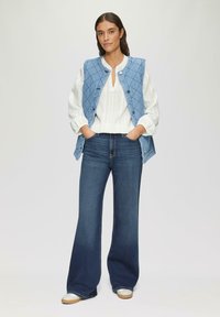 Blauwe gewatteerde bodywarmer over een wit gestreept shirt, gecombineerd met donkere flared jeans en lichte sneakers. Eenvoudige knoopdetail op de bodywarmer.