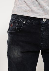 Mörk denimjeans med en blekt struktur, som har en framficka, metallknapplås och en liten brun märkestagg på sidan.