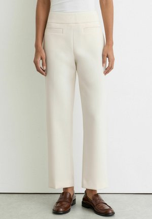 Žena oblečená v krémových nohaviciach s vysokým pásom a širokými nohavicami, v hnedých kožených mokasínach, stojaca pri bielej stene.