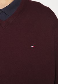 Gros plan sur une personne portant un pull col V bordeaux par-dessus une chemise à col bleu marine, avec un petit logo rouge, blanc et bleu sur la poitrine.