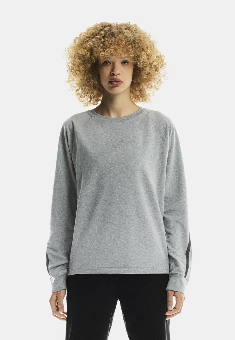 Person with curly hair wearing a light gray long-sleeve shirt and black pants, standing against a plain white background.