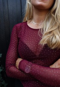 Bordowa siateczkowa bluzka z długim rękawem z srebrnymi ćwiekami, z niskim dekoltem i dopasowanymi rękawami, eksponująca szczegółowy zegarek na nadgarstku.