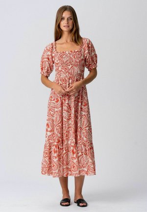 Femme debout portant une robe midi à manches bouffantes avec des motifs floraux rouges, assortie à des sandales noires à enfiler ouvertes au bout.