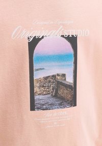 Cadre de fenêtre en pierre cintré donnant sur l'océan bleu, une côte rocheuse et un chemin pavé, avec le texte « Originals Studio » sur fond rose pâle.