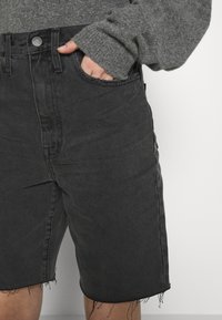 Mustat farkkushortsit, joissa on korkea vyötärö, rispaantunut helma ja nappikiinnitys. Teksturoitu kangas näyttää hienovaraista kulumista ja hieman väljää istuvuutta.