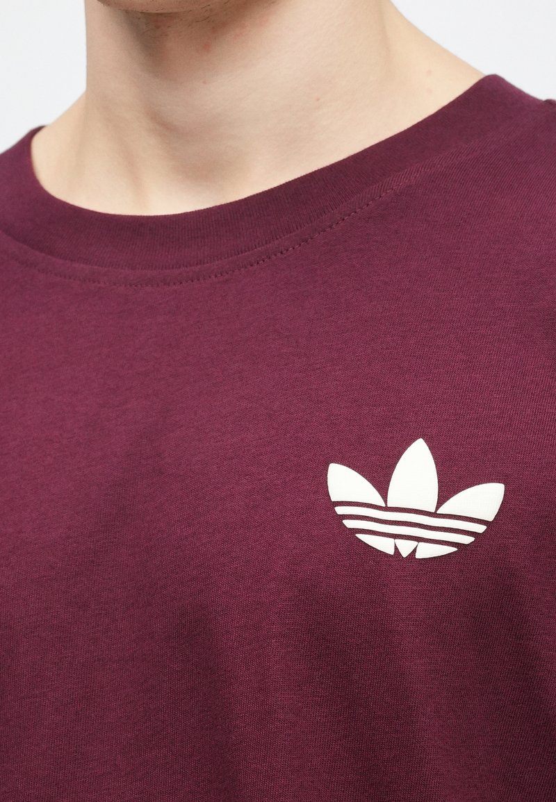 Camiseta de algodón color burdeos con un logotipo de tres rayas en blanco en el pecho. Cuello redondo con un acabado de tejido liso y texturizado.