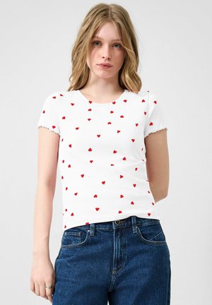 Junge Frau mit welligem blondem Haar, die ein weißes, eng anliegendes Kurzarmshirt mit kleinen roten Herzen und eine blaue hochtaillierte Jeans trägt.