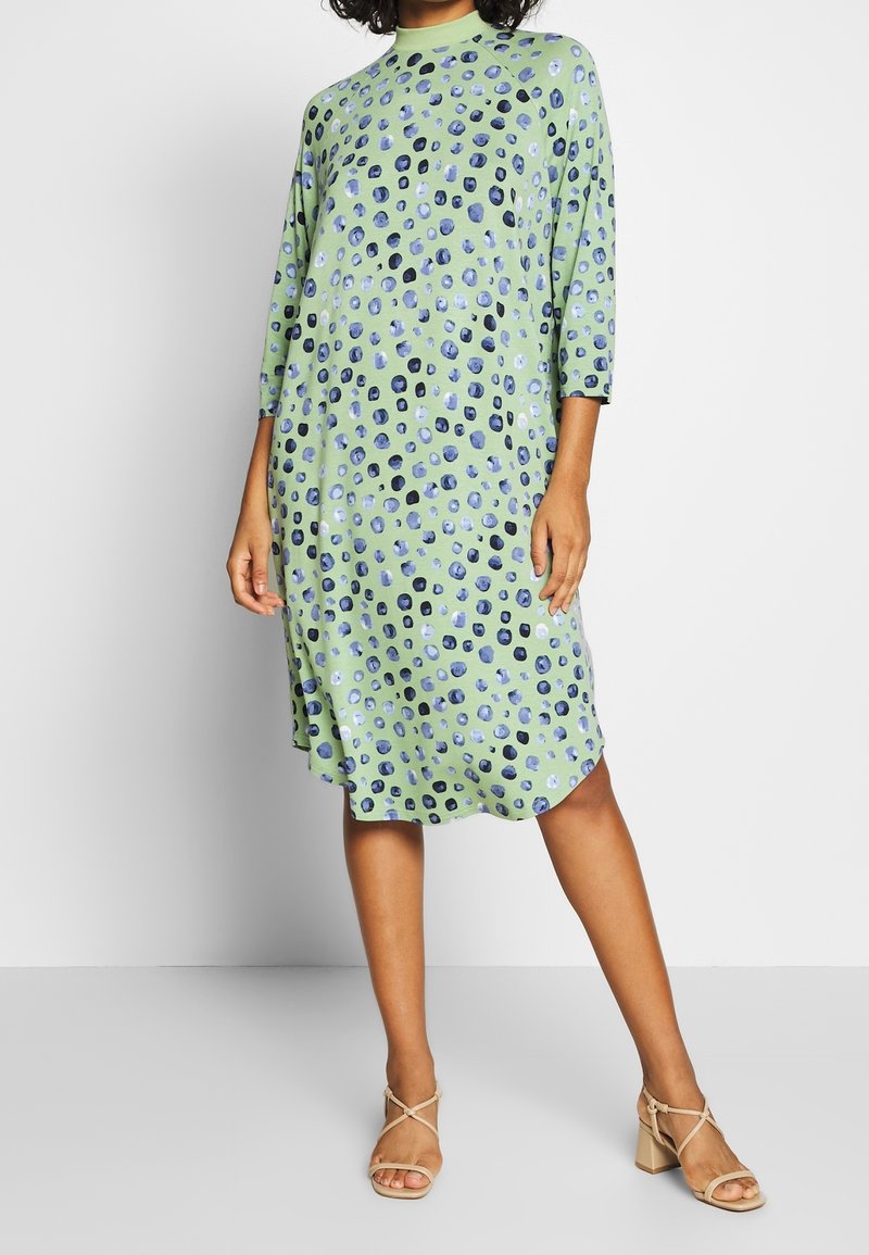 Personne portant une robe vert clair mi-longue aux genoux avec des motifs circulaires bleus et noirs et des sandales beiges à talons blocs ouvertes à l'avant.