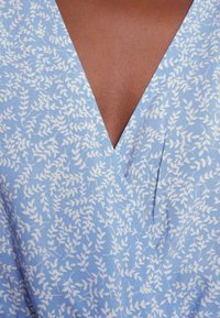 Robe à col en V en tissu bleu pâle avec motif de feuilles blanches portée par une personne à la peau brune visible au niveau du décolleté.