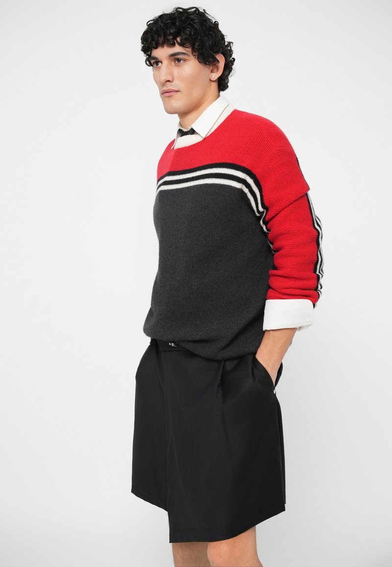 Suéter de punto a rayas rojas y grises con cuello redondo, llevado sobre una camisa blanca de cuello, combinado con shorts negros con plisado.