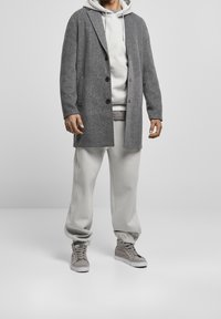 Grå herringbone-frakke med skrå lapeller, sorte knapper, båret over en lys grå hoodie og matchende sweatpants, parret med grå sneakers.