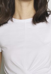 Overkroppen af en person med skulderlangt mørkt hår iført en simpel hvid T-shirt med rund hals, mod en neutral baggrund.