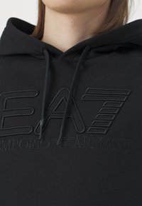 Fekete kapucnis pulóver nagy, dombornyomott "EA7" logóval és "Emporio Armani" felirattal. Kihúzható kapucnival és puha, texturált felülettel rendelkezik.