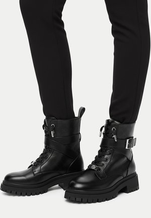 Schwarze Leder-Stiefeletten mit klobigen Sohlen, Schnürung vorne, seitlichen Schnallenriemen und Zuglaschen, getragen zu schwarzen Slim-Hosen.