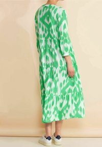 Pitkä vihreä mekko, jossa on abstrakti valkoinen kuvio, kolme kerrosta, 3/4-hihat ja hieman levenevä helma, yhdistettynä valkoisiin sneaker-kengiin.