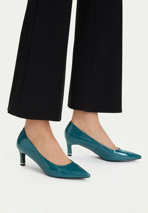 Zapatos de charol verde azulado con punta estrecha y tacón medio, usados con pantalones negros anchos y recortados sobre un fondo claro.