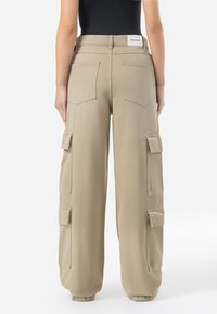 Beige cargo bukser med løs pasform, med sidelommer, elastiske mansjetter og en etikett på livet. Stoff i bomullsblanding med en glatt tekstur.