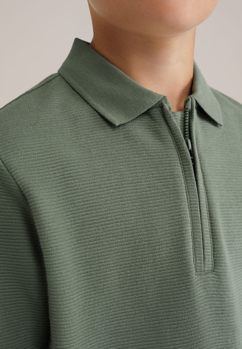 Zelená polo košile na zip s texturovanou látkou, krátkým límcem a centrálním zipem. Design je jednoduchý a neformální, vhodný pro každodenní nošení.