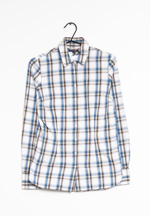 Chemise à manches longues boutonnée avec motif à carreaux bleu, marron et blanc accrochée sur un cintre noir sur fond blanc.