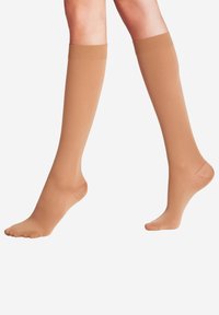 Gambe con calze compressive beige al ginocchio su uno sfondo bianco, un piede piatto e un dito del piede puntato verso il basso.