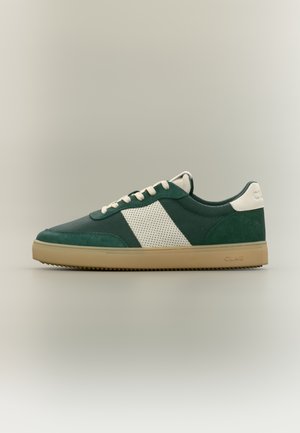 Pantof sport verde închis cu dunga laterală din plasă albă, șireturi crem, talpă bej și etichetă albă la călcâi pe fundal neutru.