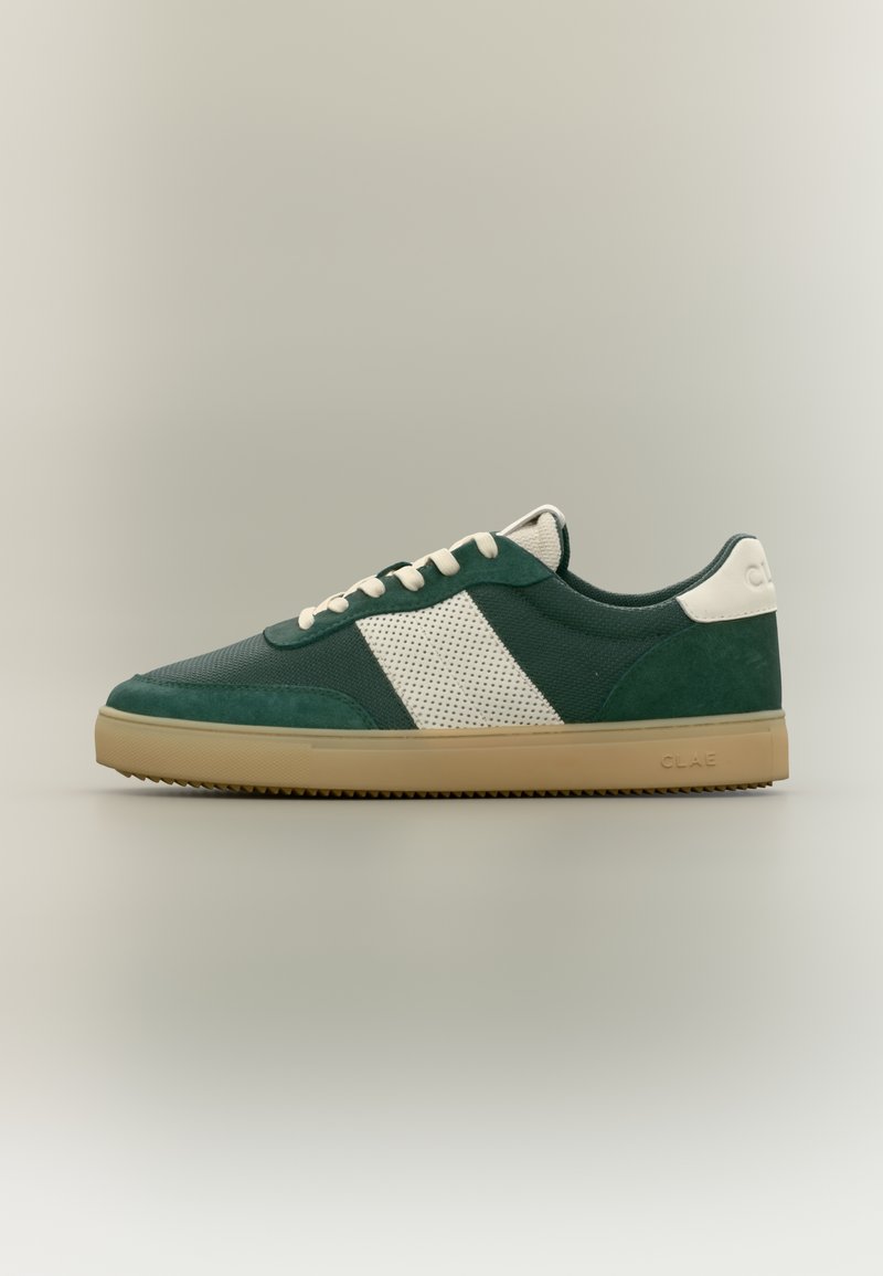 Σκούρο πράσινο αθλητικό παπούτσι με λευκή ρίγα διχτυού στο πλάι, κρεμ κορδόνια, μπεζ σόλα και λευκή λωρίδα στη φτέρνα σε ουδέτερο φόντο.