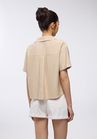 Blusa beige a maniche corte con tessuto testurizzato, colletto e vestibilità rilassata, abbinata a pantaloni corti bianchi con risvolto.